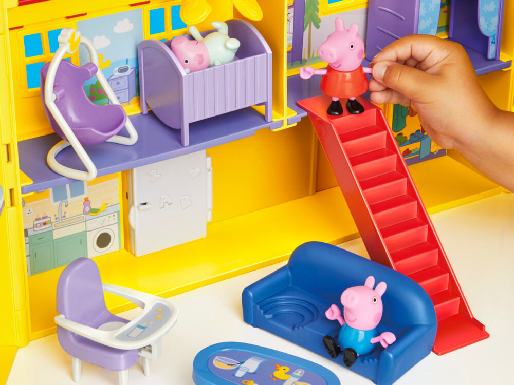 Zestaw figurek HASBRO Świnka Peppa Duży dom rodzinny Peppy G05085L0 Widok na wnętrze rozłożonego domku świnki Peppy podczas zabawy. Dziecko ustawia figurkę Peppy na szczycie czerwonych schodów. Na parterze widać niebieską kanapę z figurką George'a, a na piętrze fioletowe łóżeczko i huśtawkę.