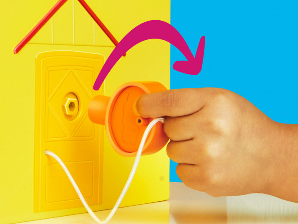 Zestaw figurek HASBRO Świnka Peppa Duży dom rodzinny Peppy G05085L0 Zbliżenie na żółtą ścianę zewnętrzną domku Peppa Pig. Dłoń dziecka wkłada pomarańczowy klucz do otworu w drzwiach. Fioletowa strzałka pokazuje kierunek obrotu klucza, sugerując mechanizm otwierania lub aktywacji dźwięku.