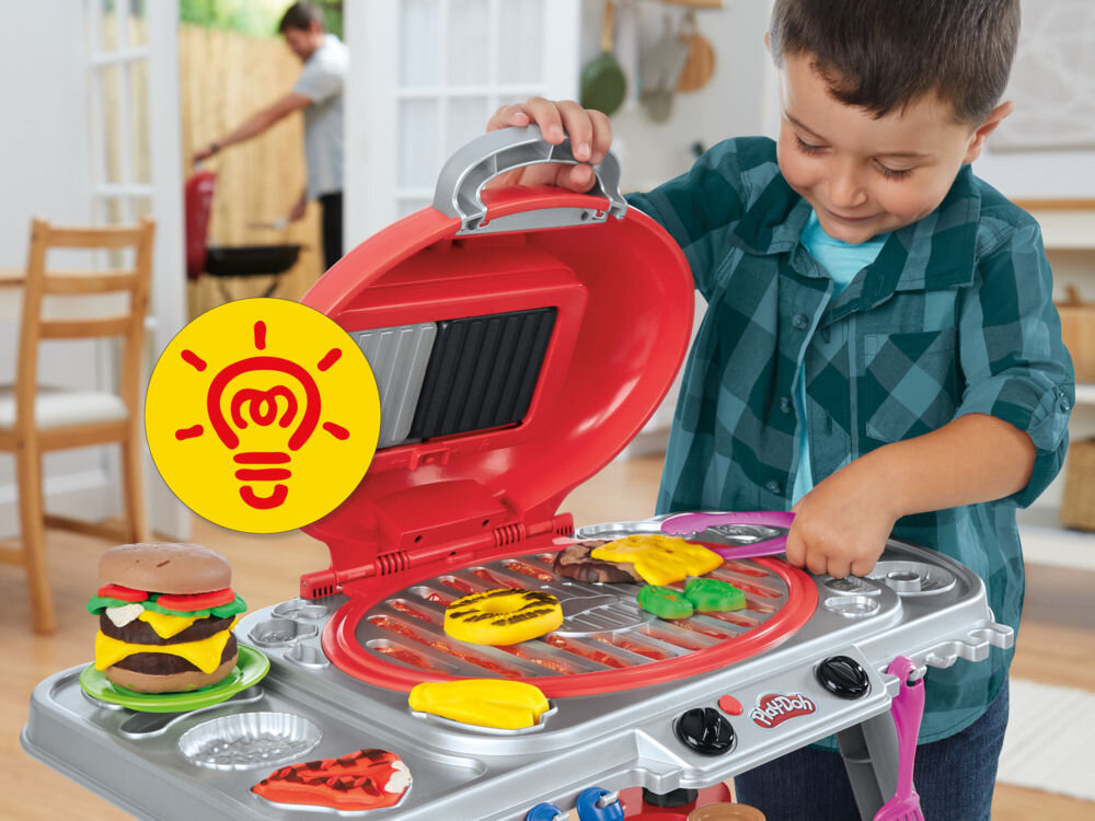 Ciastolina PLAY-DOH Skwierczący grill G05005L0 Chłopiec bawi się zestawem Play-Doh Skwierczący Grill, podnosząc czerwoną pokrywę. Używa różowej szpatułki do przewracania ciastolinowych burgerów i dodatków na ruszcie. Obok stoi gotowy, wielowarstwowy burger z ciastoliny.