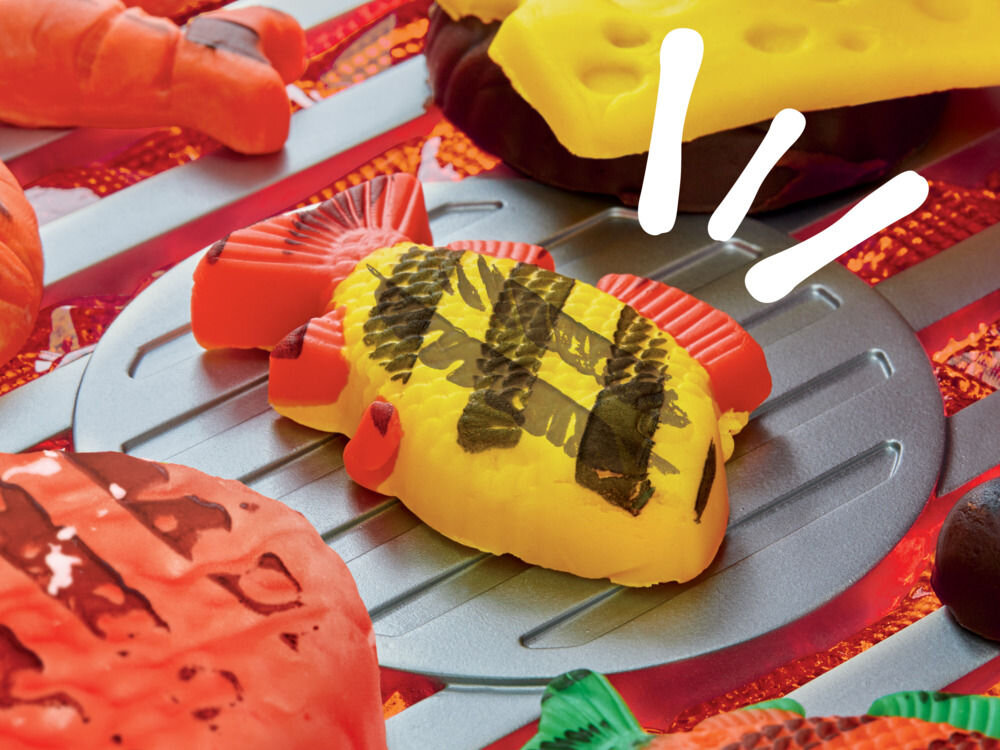 Ciastolina PLAY-DOH Skwierczący grill G05005L0 Bardzo bliskie ujęcie ciastolinowej żółtej ryby z wyraźnymi śladami grillowania (czarne prążki), leżącej na srebrnej łopatce. Prezentacja efektu formowania z zestawu Play-Doh Skwierczący Grill.