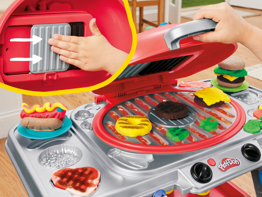 Ciastolina PLAY-DOH Skwierczący grill G05005L0 Zestaw Play-Doh Grill w akcji. Zbliżenie pokazuje, jak dziecko umieszcza wyjmowaną praskę (formę) w pokrywie grilla, aby odciskać ślady rusztu na cieście. Na płycie głównej widoczny hot dog i uformowane burgery.