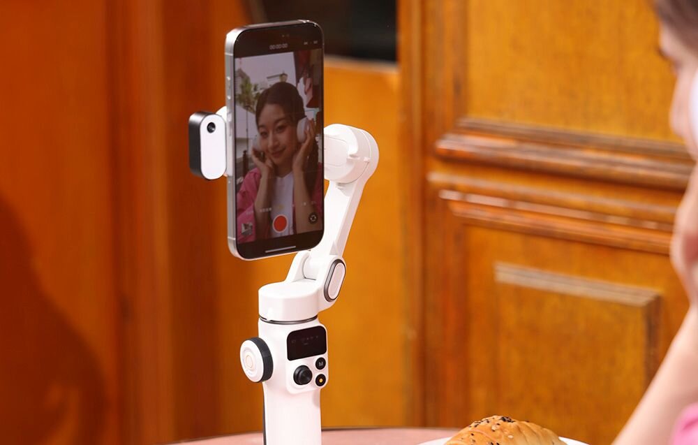 Młoda kobieta z zaplecionymi warkoczami, ubrana w różową koszulę, idzie ulicą i trzyma przed sobą biały gimbal ze smartfonem, nagrywając wideo
