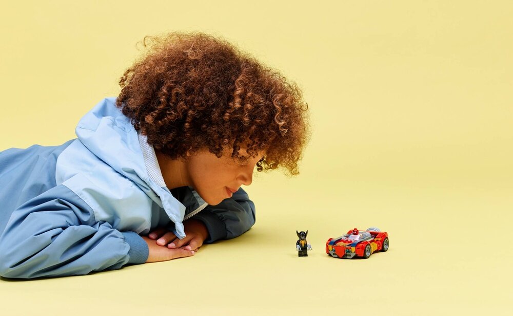 Osoba w niebieskiej kurtce patrzy na zestaw LEGO z figurką i czerwonym samochodem na żółtym tle.