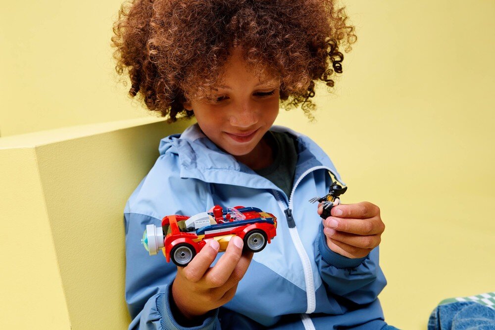 Dziecko w niebieskiej kurtce trzyma w rękach zabawkowy samochód i figurkę. Tło jest jednolite, w kolorze żółtym.