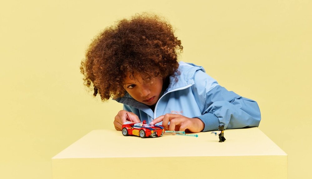 Dziecko w niebieskiej kurtce bawi się zestawem LEGO, który zawiera czerwony samochód i małą figurkę. Tło jest jednolite, w kolorze żółtym.