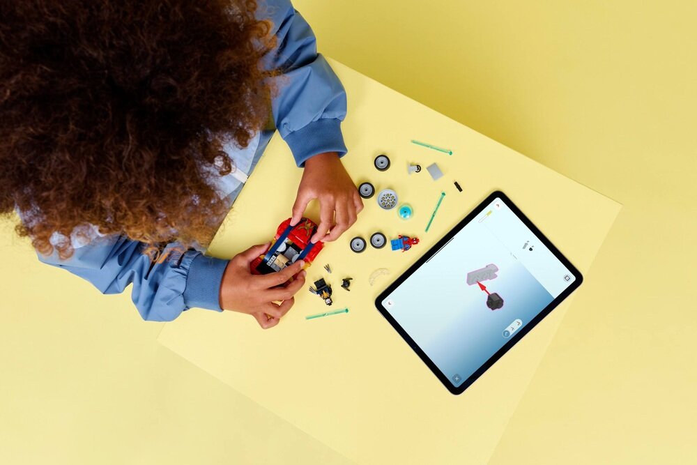 Osoba składa zestaw klocków LEGO na żółtym stole, obok leży tablet z instrukcją. Na stole widoczne są różne elementy LEGO, w tym koła i figurki.