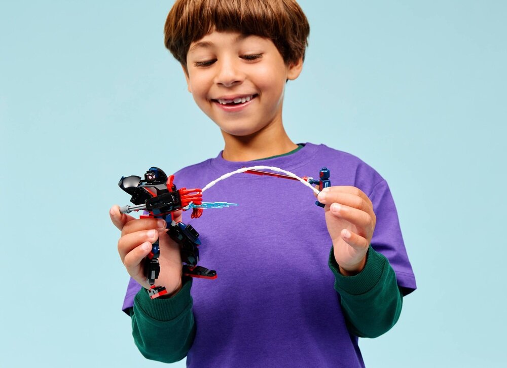 Dziecko w fioletowej koszulce trzyma w rękach zestaw klocków LEGO przedstawiający dwie postacie z serii Marvel. Tło jest jednolite, w jasnym kolorze.