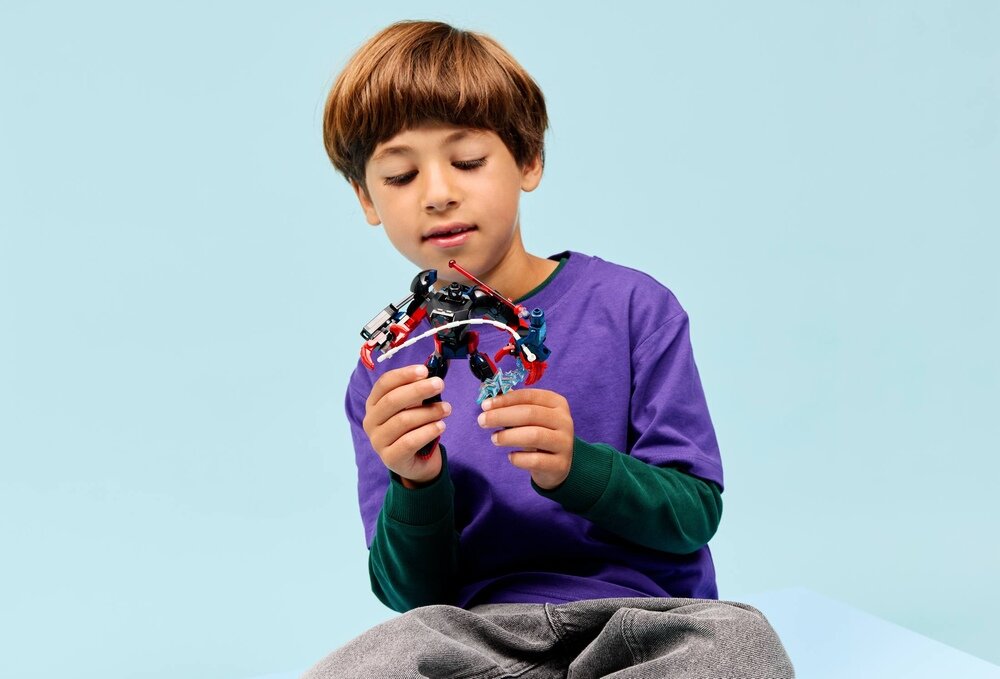 Dziecko w fioletowej koszulce trzyma zestaw LEGO z figurkami i elementami konstrukcyjnymi. Tło jest jednolite, jasnoniebieskie.