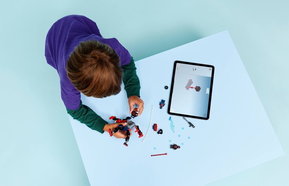 Dziecko bawi się zestawem klocków LEGO na białym stole. Obok znajduje się tablet z wyświetlonym obrazem klocków LEGO.