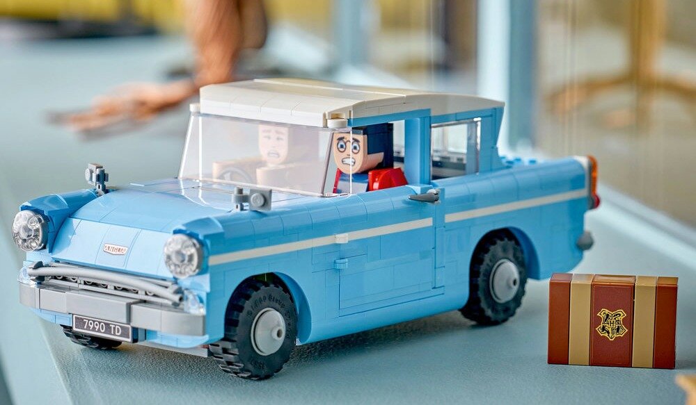 Model samochodu LEGO w kolorze niebieskim z białym dachem, z dwoma figurkami w środku. Obok samochodu znajduje się mała brązowa skrzynka z herbem Hogwartu.