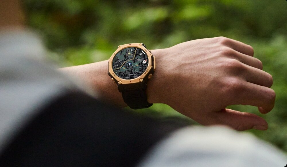 Osoba nosi smartwatch z okrągłą tarczą i czarnym paskiem, wyświetlający szczegółowy wzór na ekranie. W tle widoczna jest rozmyta zieleń, sugerująca otoczenie na świeżym powietrzu.