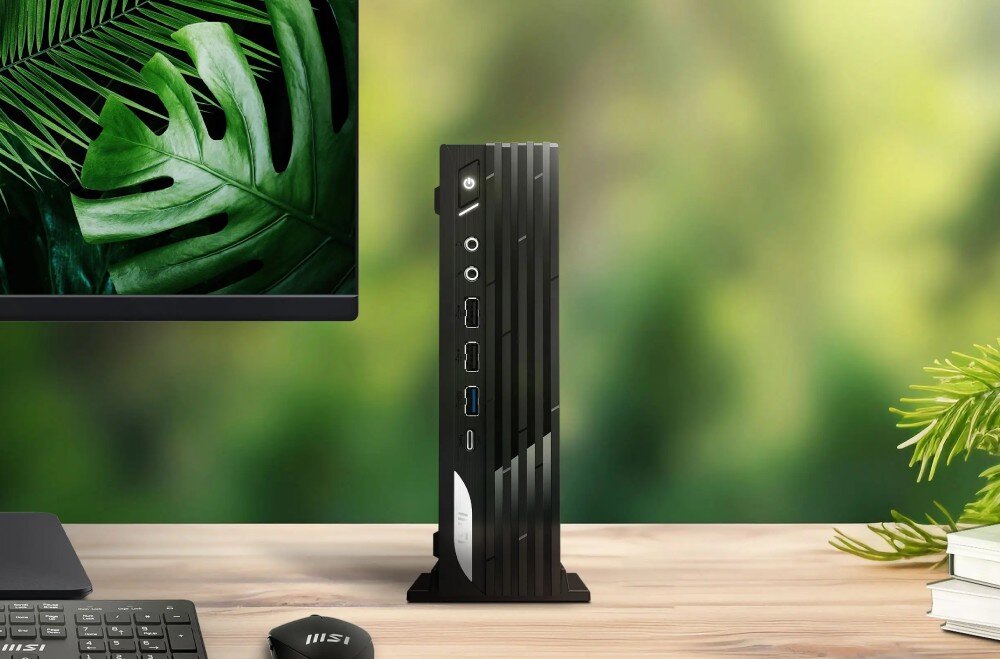 Komputer MSI Pro DP21 14M-889EU Czarny komputer stacjonarny typu mini PC stojący pionowo na drewnianym biurku obok monitora wyświetlającego zielony liść, w tle rozmyte zielone rośliny, opis