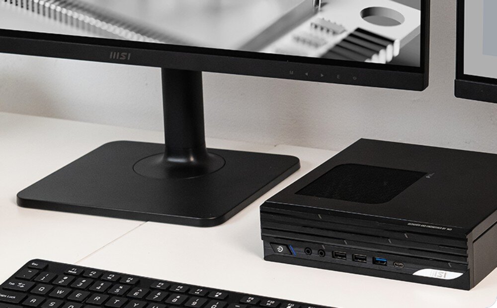 Komputer MSI Pro DP21 14M-889EU Komputer typu mini PC leżący poziomo na białym biurku obok podstawy czarnego monitora oraz fragmentu czarnej klawiatury, wytrzymałość, testy