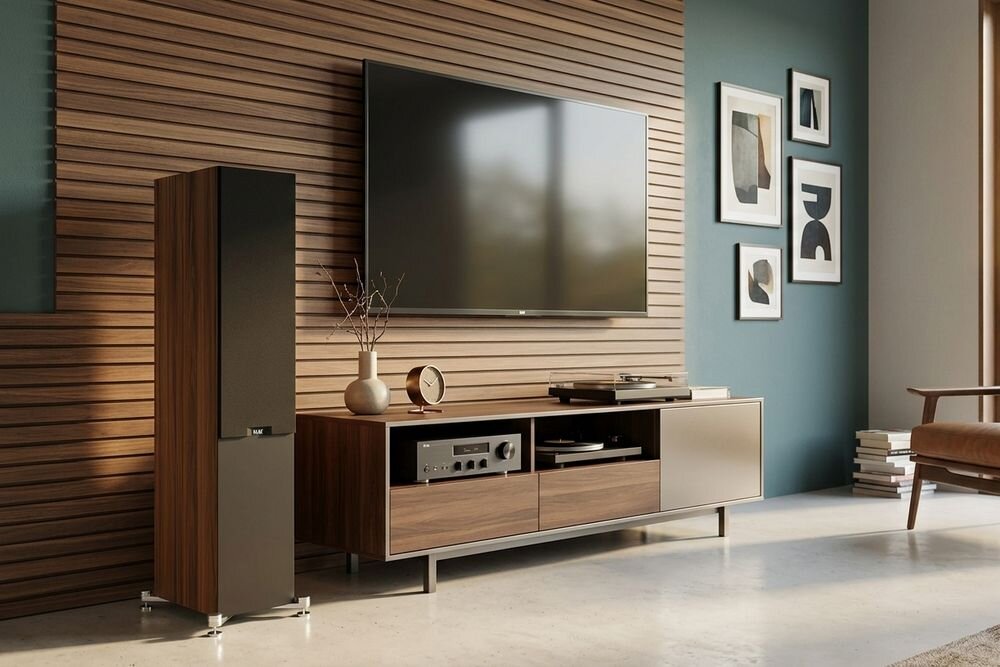 W nowoczesnym salonie znajduje się duży telewizor zamontowany na ścianie z drewnianymi panelami oraz kolumna głośnikowa w kolorze orzechowym. Obok telewizora stoi szafka RTV z różnymi urządzeniami elektronicznymi i dekoracjami.