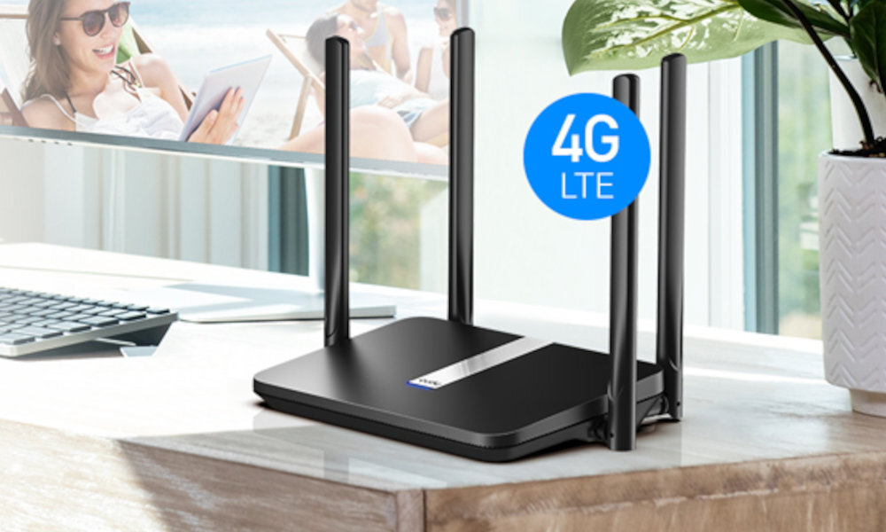Na drewnianym biurku stoi czarny router z czterema antenami, obok którego widnieje niebieska ikona z napisem \'4G LTE\'. W tle widać fragment okna i roślinę w białej doniczce.