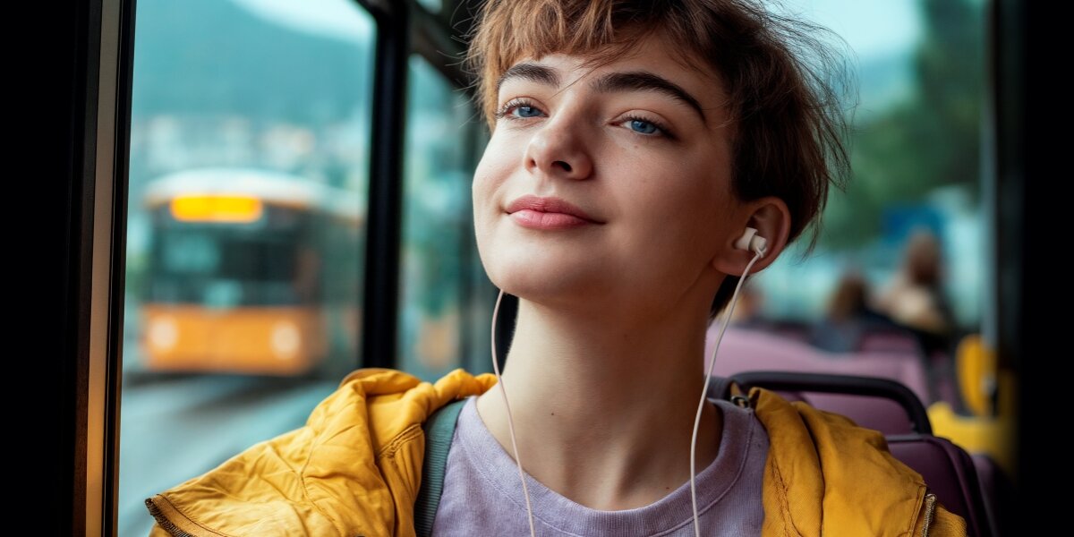 Młoda kobieta w żółtej kurtce i fioletowej koszulce, słuchająca muzyki przez białe słuchawki douszne Hama w autobusie. Scena podróży, w tle widać zamazany widok ulicy i inny autobus.