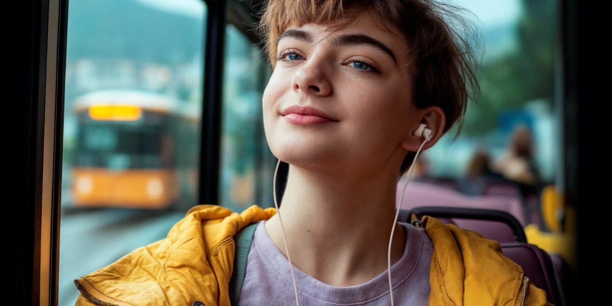 Młoda kobieta w żółtej kurtce, słuchająca muzyki przez białe słuchawki douszne Hama z wtyczką USB-C, siedząc w autobusie. Kobieta ma zrelaksowany wyraz twarzy, w tle widać ulicę i inny autobus.