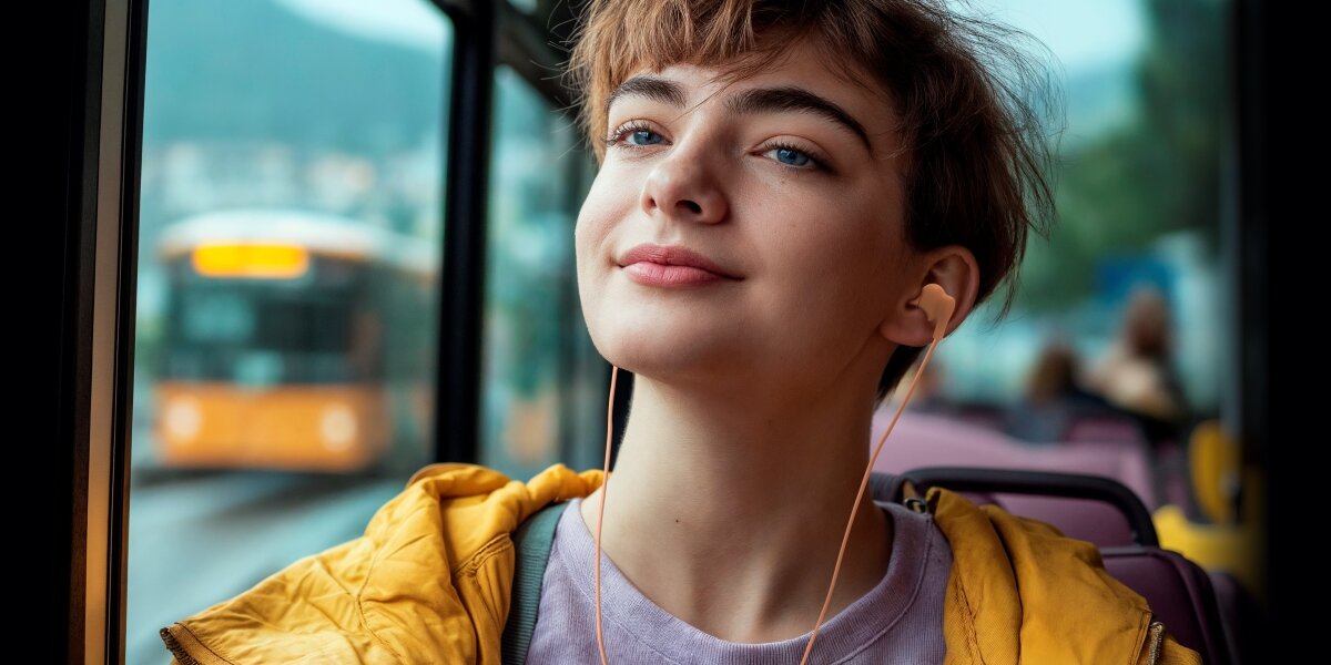 Młoda kobieta w żółtej kurtce, słuchająca muzyki przez brzoskwiniowe/różowe słuchawki douszne Hama z wtyczką USB-C, siedząc w autobusie. Kobieta ma zrelaksowany wyraz twarzy, w tle widać ulicę i inny autobus.