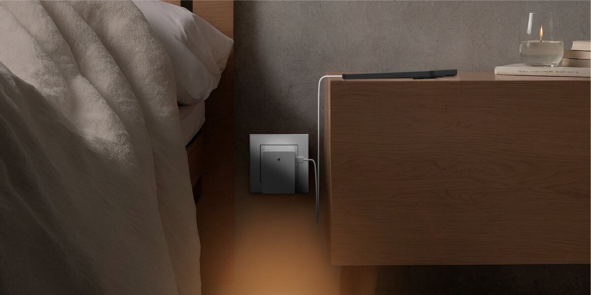 Lampka nocna HAMA 223506 z czujnikiem zmierzchu Lampka nocna Hama wpięta do gniazdka przy łóżku, ładująca smartfona leżącego na drewnianym stoliku nocnym.