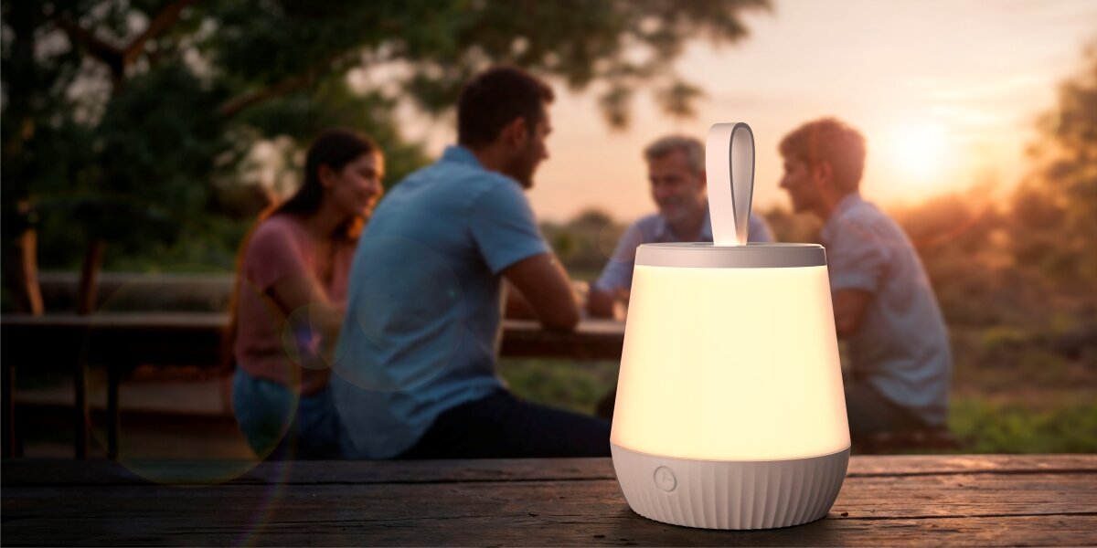 Lampka nocna HAMA Mobile Przenośna lampka ogrodowa LED z uchwytem, świecąca ciepłym światłem na drewnianym stole podczas wieczornego spotkania grupy przyjaciół w ogrodzie.