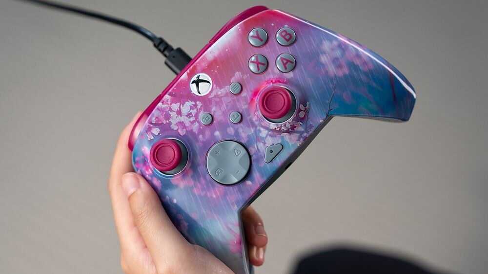 Kontroler TURTLE BEACH Xbox Rematch Advanced Kyoto Bloom TBC-2404-45 Zbliżenie na pad Kyoto Bloom trzymany w jednej dłoni, pokazujące detale kwiecistego nadruku oraz różowe drążki analogowe kontrola różnych aspektów ustawienia audio jeden przycisk szybkie włączenie lub wyłączenie mikrofonu regulacja czułości spustów bezpłatna aplikacja Control Hub konsole Xbox PC