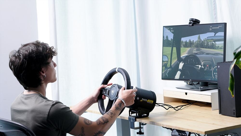 Kierownica Turtle Beach Velocity OneRace KD3 Użytkownik siedzący przy biurku przed monitorem, korzystający z zestawu kierownicy i bazy Turtle Beach VelocityOne Race układ Force Feedback pięć różnych profili ustawień aplikacja VelocityOne Tuner dostosowanie czułości regulacja oświetlenia