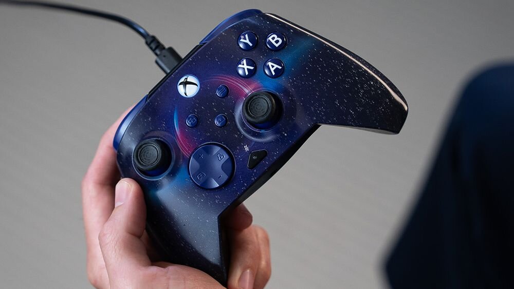 Kontroler TURTLE BEACH Xbox Rematch Advanced Dark Cosmos TBC-2404-05 Zbliżenie na pad Dark Cosmos trzymany w jednej dłoni, pokazujące detale gwiezdnego nadruku na obudowie kontrola różnych aspektów ustawienia audio jeden przycisk szybkie włączenie lub wyłączenie mikrofonu regulacja czułości spustów bezpłatna aplikacja Control Hub konsole Xbox PC