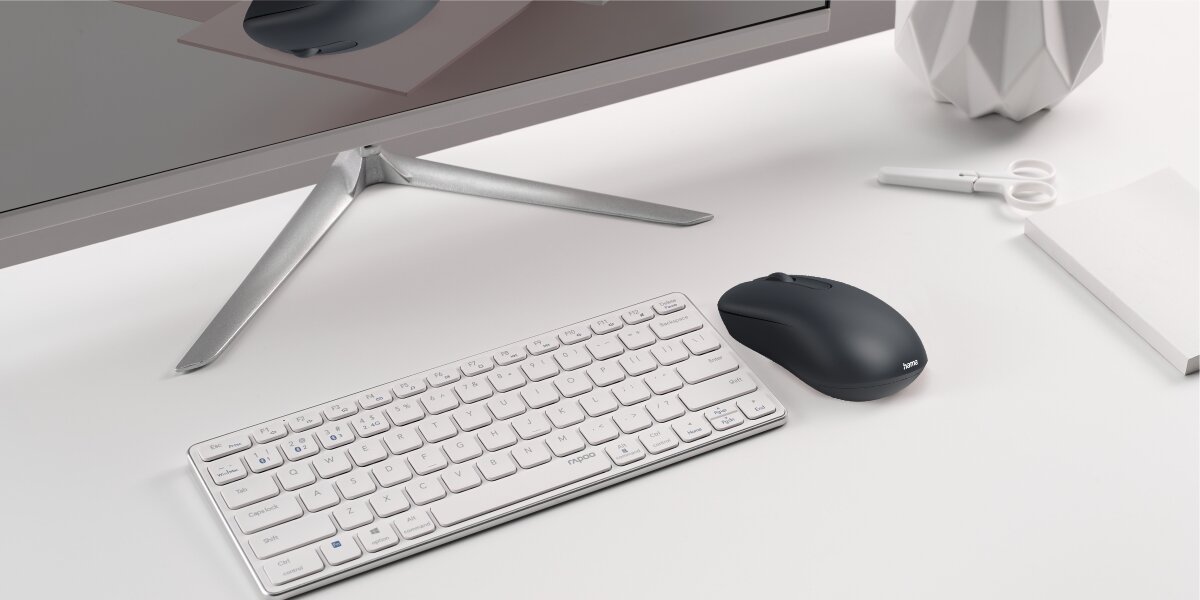 Czarna mysz bezprzewodowa na biurku obok białej, kompaktowej klawiatury i monitora, w eleganckim, biurowym otoczeniu.