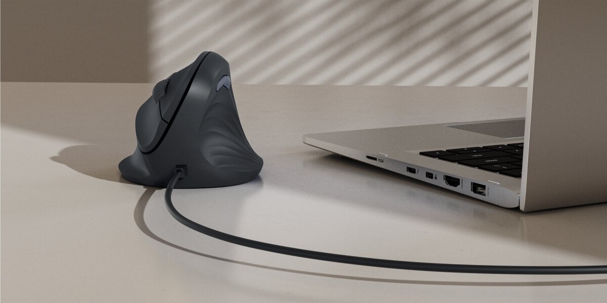 Pionowa mysz ergonomiczna, podłączona kablem USB do laptopa na biurku, z widocznym profilowanym kształtem, minimalizującym napięcie nadgarstka.