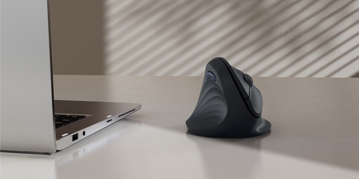 Pionowa mysz ergonomiczna HAMA na biurku, stojąca obok otwartego laptopa. W tle widać cień żaluzji.