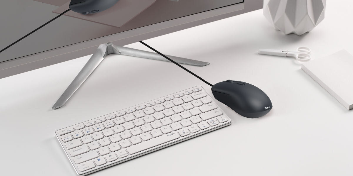 Komfort dla każdej dłoni Czarna myszka przewodowa leżąca na białym biurku obok białej, kompaktowej klawiatury bezprzewodowej, podłączona do monitora, co ukazuje typowe środowisko pracy.