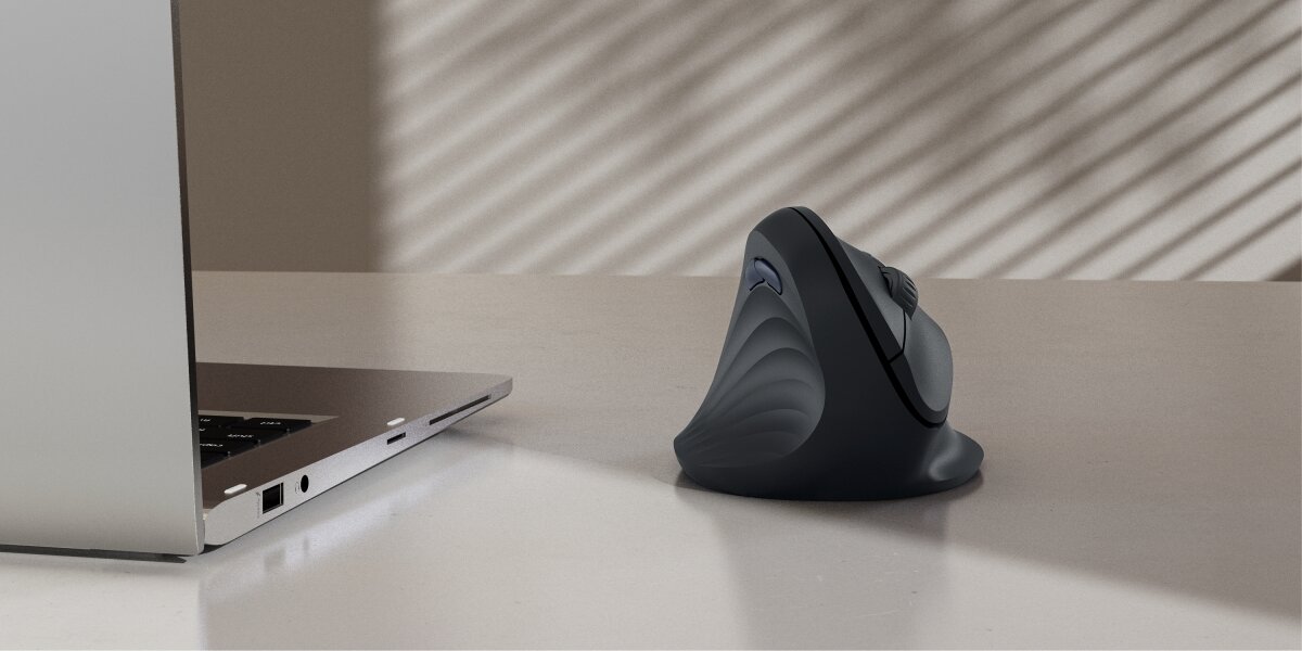 Pionowa mysz ergonomiczna Hama z falistą teksturą uchwytu, ustawiona na białym biurku obok otwartego laptopa. W tle widać cień rzucany przez żaluzje.