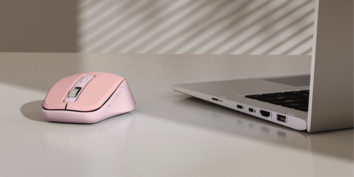 Ergonomiczna mysz bezprzewodowa w kolorze różowym (pastelowy róż) leżąca na biurku obok srebrnego laptopa, prezentująca nowoczesne, kolorowe stanowisko pracy.