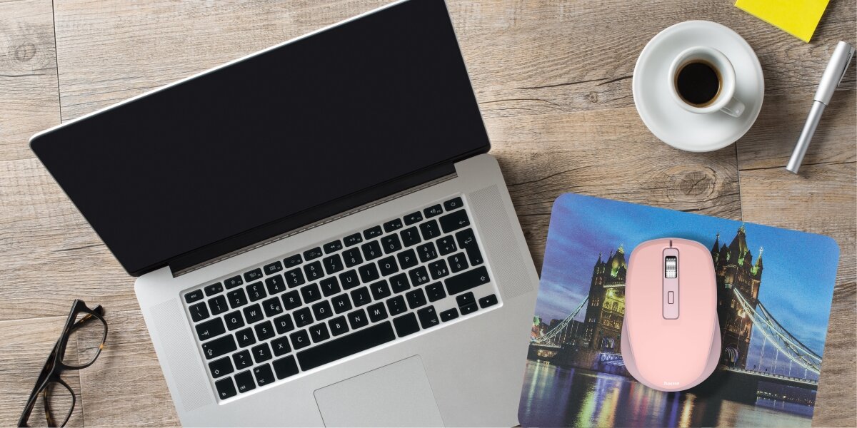 Różowa mysz bezprzewodowa na podkładce ze wzorem mostu Tower Bridge w Londynie, leżąca obok otwartego laptopa i kawy.