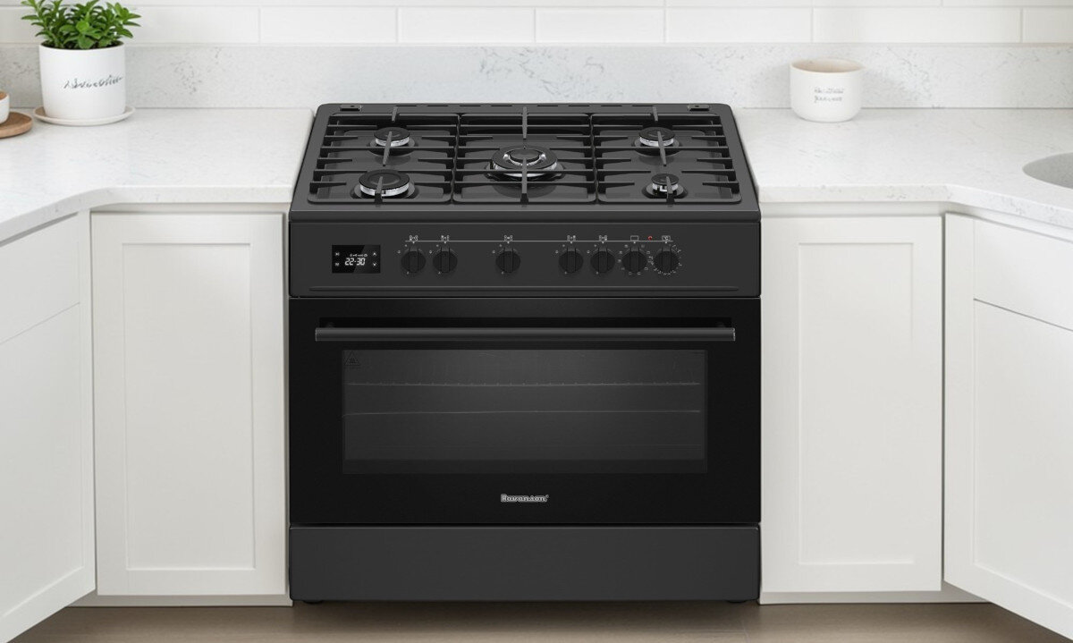 Czarna kuchenka gazowa z czterema palnikami i piekarnikiem, umieszczona pomiędzy białymi szafkami kuchennymi. Na froncie kuchenki widoczne jest logo \'RAVANSON\'.
