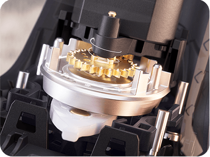 Zbliżenie na mechanizm wewnętrzny urządzenia z widocznymi metalowymi i plastikowymi elementami, w tym złotymi zębatkami.