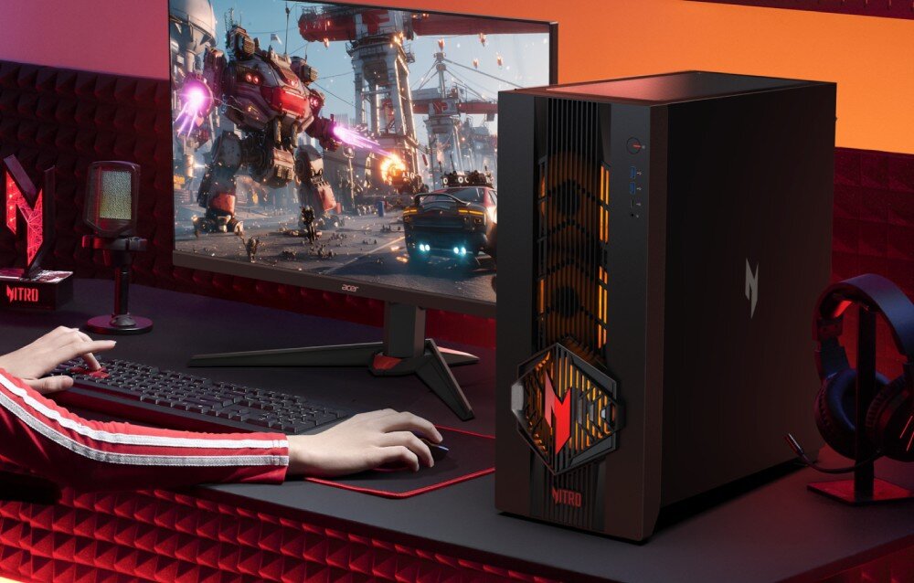 Na biurku znajduje się komputer stacjonarny z logo \'Nitro\' oraz monitor wyświetlający scenę z gry z robotami. Obok klawiatury i myszki widoczny jest mikrofon i słuchawki.