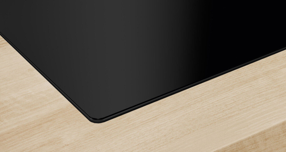 PŁYTA BOSCH PXL811H26E Na zdjęciu widać zbliżenie narożnika czarnej, szklanej płyty grzewczej osadzonej w jasnym, drewnianym blacie. Gładka krawędź szkła podkreśla precyzyjne wykończenie i estetyczne dopasowanie do blatu komfort gotowania estetyka bezramkowa minimalistyczny styl łatwe czyszczenie aranżacja kuchni