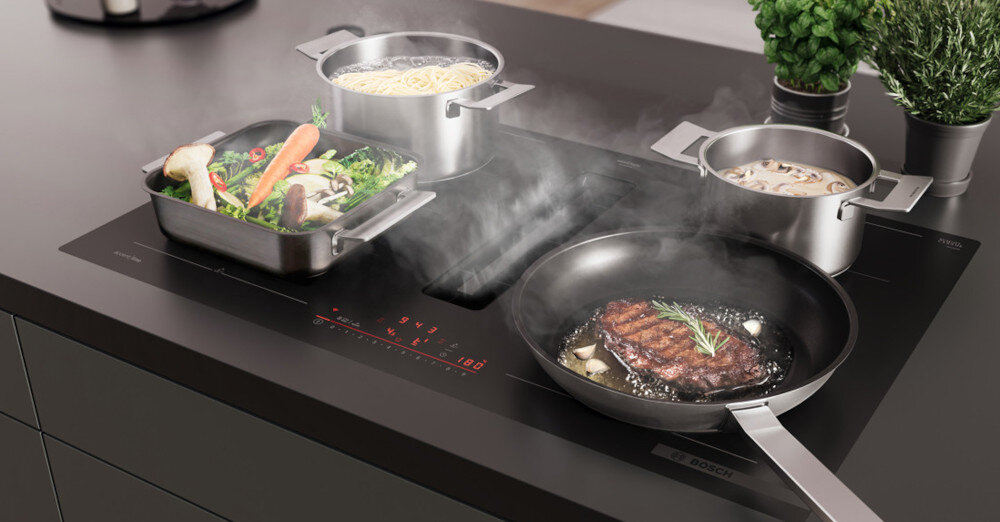 PŁYTA BOSCH PXL811H26E Na zdjęciu widać płytę indukcyjną Bosch zintegrowaną z wyciągiem środkowym, na której jednocześnie gotują się warzywa w brytfannie, makaron w garnku oraz sos w mniejszym naczyniu, a na patelni smaży się stek. Para z naczyń jest od razu zasysana do centralnego wlotu, co podkreśla skuteczne odprowadzanie oparów bez tradycyjnego okapu QuickStart wykrywanie naczynia automatyczna strefa grzania panel sterowania szybkie gotowanie poziom grzania