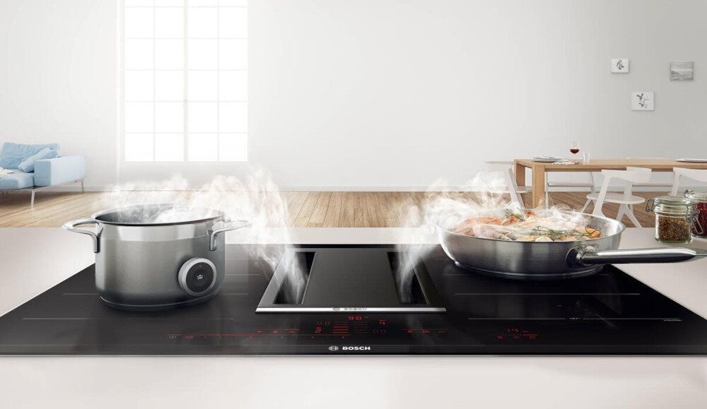 PŁYTA BOSCH PXL811H26E Na zdjęciu widoczna jest czarna płyta zintegrowana z wyciągiem środkowym, na której stoją dwa naczynia z gotującą się potrawą i unoszącą się parą. W tle widać otwartą przestrzeń dzienną z salonem i jadalnią, co podkreśla, że odciąg pary odbywa się bez tradycyjnego okapu nad płytą Zintegrowany wyciąg płyta grzewcza wyciąg indukcja elegancja odciąg świeże powietrze przestrzeń funkcjonalność design wygoda
