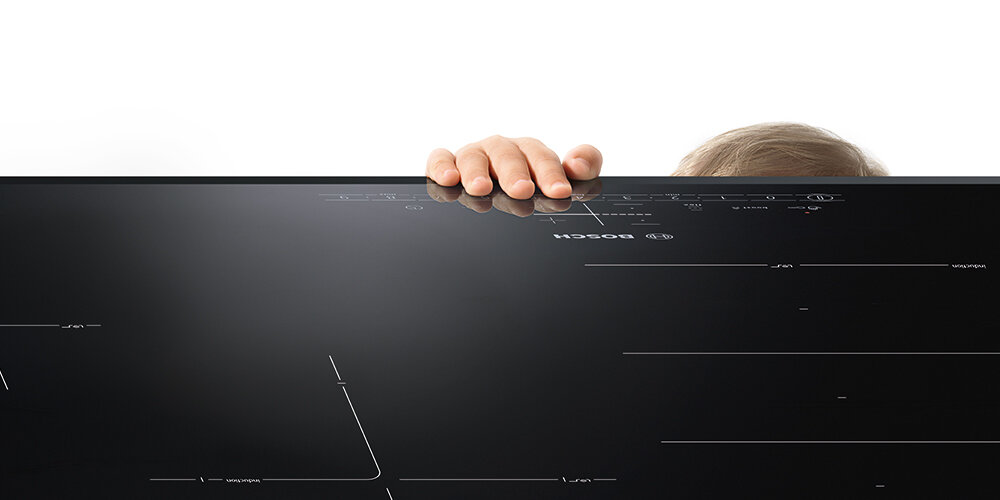 PŁYTA BOSCH PXL811H26E Na zdjęciu widać dużą, czarną płytę indukcyjną Bosch, za której krawędź chwyta ręką małe dziecko, ledwo wystające znad blatu. Ujęcie podkreśla rozmiar płyty oraz nawiązuje do funkcji bezpieczeństwa chroniących najmłodszych użytkowników zabezpieczenie przed dziećmi blokada panelu bezpieczeństwo spokój pewność gotowanie