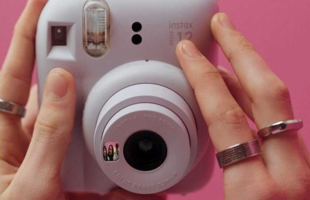 Osoba trzyma biały aparat fotograficzny z napisem \'instax mini 12\' na różowym tle. Aparat ma widoczny obiektyw i lampę błyskową.
