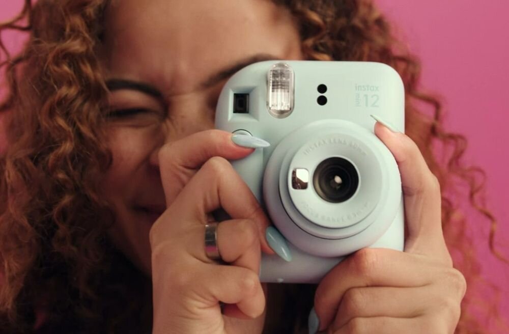 Osoba trzyma aparat fotograficzny w kolorze jasnoniebieskim z widocznym napisem \'instax mini 12\'.