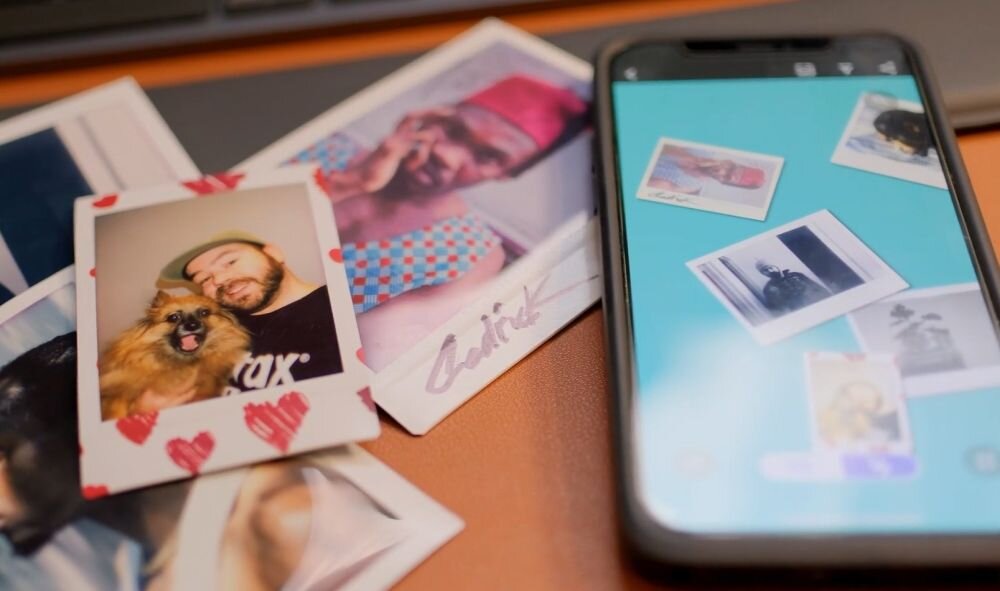 Na zdjęciu widać kilka kolorowych zdjęć natychmiastowych rozłożonych na powierzchni oraz smartfon z wyświetlonymi zdjęciami na ekranie. Jedno ze zdjęć przedstawia osobę z psem, a inne osoby w różnych pozach.