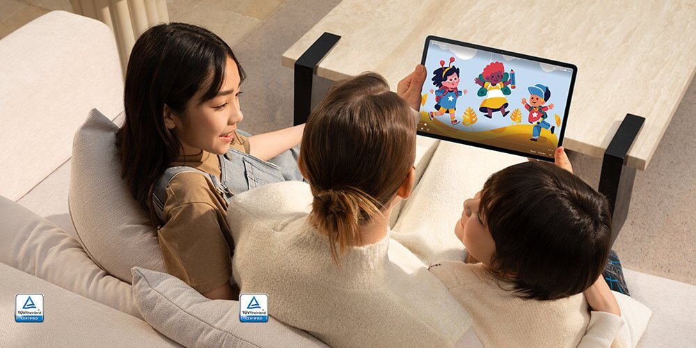 Trzy osoby siedzą na kanapie, trzymając tablet z kolorową grafiką przedstawiającą postacie z kreskówki. Na dole obrazu widoczne są dwa logotypy TÜV Rheinland.