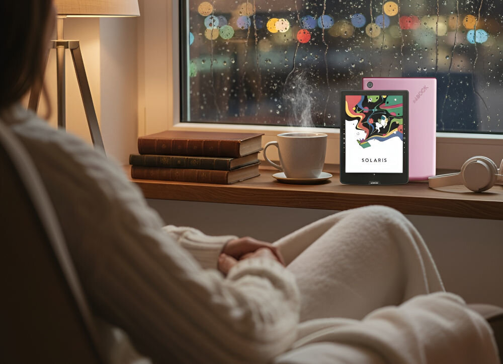 Czytnik e-booków INKBOOK Solaris Color 6 cali, Podświetlany ekran, Wi-Fi Różowy prezentacja czytnika w pokoju na parapecie obok książki kubek lampka przed kobieta siedząca na fotelu