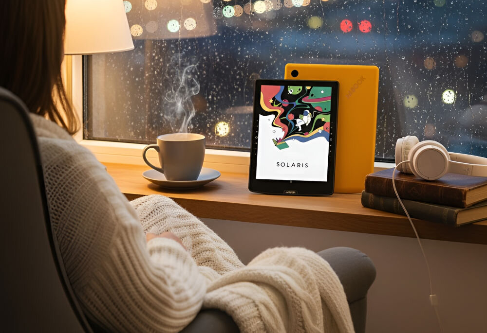 Czytnik e-booków INKBOOK Solaris Color 6 cali, Podświetlany ekran, Wi-Fi Żółty prezentacja czytnika w pokoju na parapecie obok książki kubek lampka przed kobieta siedząca na fotelu