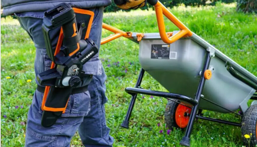 Piła akumulatorowa WORX Nitro WG325E ogród trawnik osoba niesie piłę w pokrowcu umieszczonym na nodze osoba pcha przed sobą taczkę do precyzyjnych prac pielęgnacyjnych w ogrodzie drobnych zadań wokół domu zasilanie akumulatorem 20 V usprawnia poruszanie się
