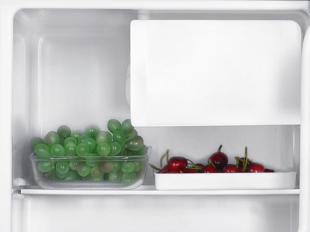 Lodówka GOTZE&JENSEN RB150K 51.5 cm Zbliżenie na górną półkę lodówki z pojemnikami pełnymi zielonych winogron i czerwonych czereśni pod komorą niskiej temperatury. 