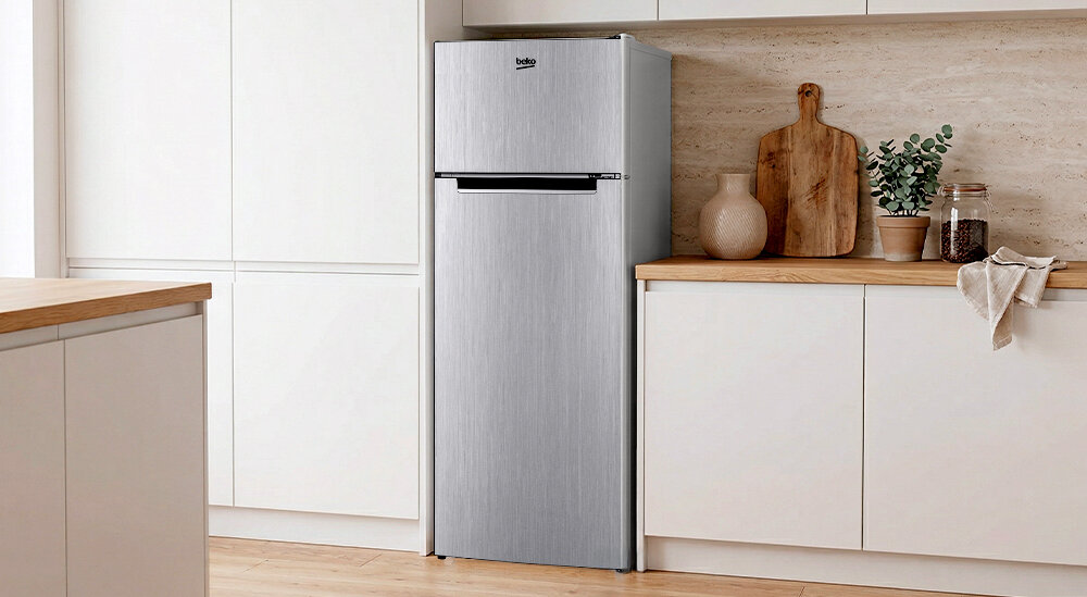 Srebrna lodówka z logo \'beko\' stoi w kuchni z jasnymi szafkami i drewnianym blatem. Obok lodówki znajdują się dekoracyjne naczynia i deska do krojenia.